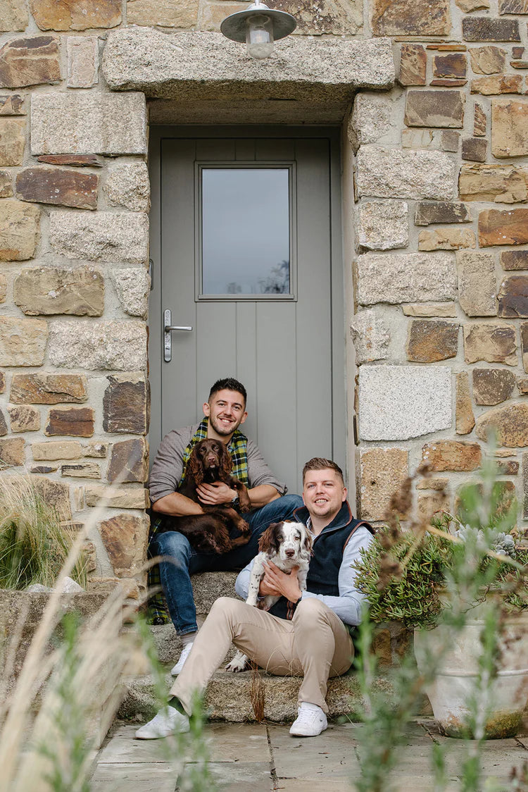 Tom and James sit on a set of steps, smiling, with dogs in their arms. The scene is outdoors, and the dogs appear content. Tom holds a small brown dog, while James cradles a larger, spotted dog. The background includes a set of steps, suggesting an outdoor setting.
