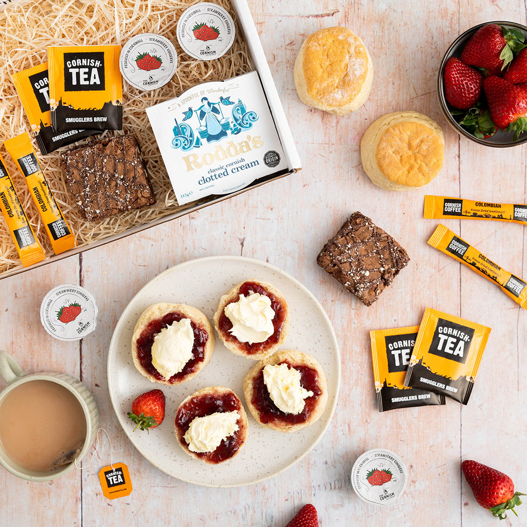 The contents of a cream tea hamper with extra coffee sachets and two brownies, packaged in a branded box on a wooden floor.