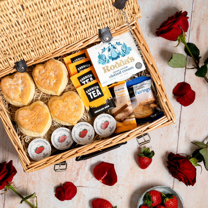 A cream tea hamper with love heart scones in a wicker basket. The hamper is placed on a wooden floor with roses laid out.