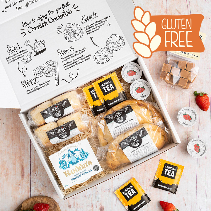 A hamper comprising of gluten free pasties and scones, tea, jam, cream and fudge. The hamper is packaged in a branded box on a wooden floor
